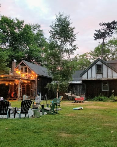 New England cottage at dusk with string lights and Adirondack chairs on a wide lawn surrounded by trees