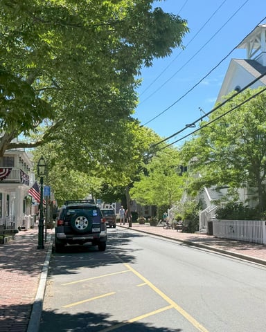 Shingle-style main street with brick sidewalk, summer trees, and white clapboard buildings on a sunny Cape Cod day