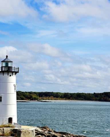 White lighthouse standing at the edge of a calm harbor with a green wooded shoreline in the background