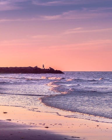 Lone fisherman silhouetted on a jetty at pink and purple dusk over the ocean