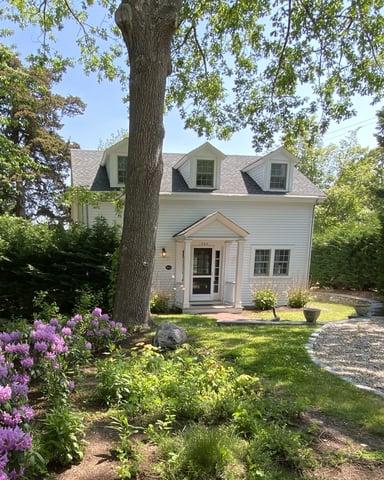 White cottage with dormers surrounded by mature trees, purple rhododendrons, and gravel driveway