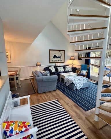 Open-plan living room with white spiral staircase, navy striped rug, sofa, armchair, and glass doors opening to deck with ocean view.