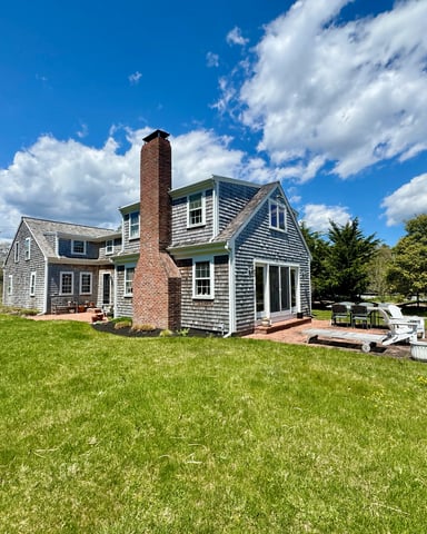Two-story shingled house with brick chimney and rear patio on large grassy lawn under blue sky