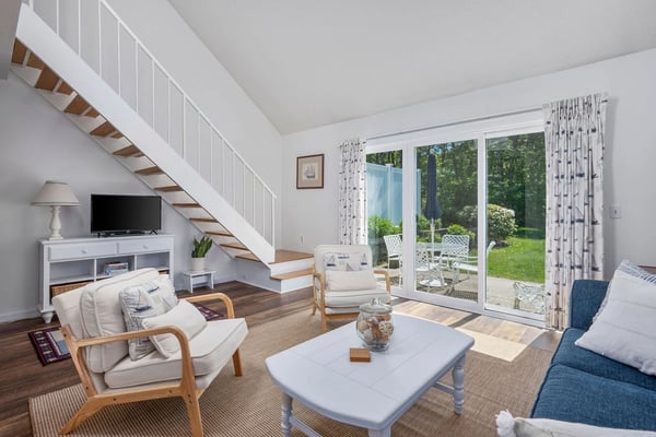 Bright Cape Cod living room with straight staircase, sailboat-print curtains, and garden patio doors