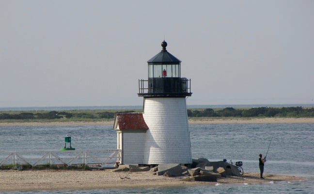 White Cape Cod lighthouse on a sandy spit with a lone fisherman casting into calm water