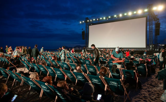 Outdoor cinema by the sea at night with rows of deck chairs facing a large screen
